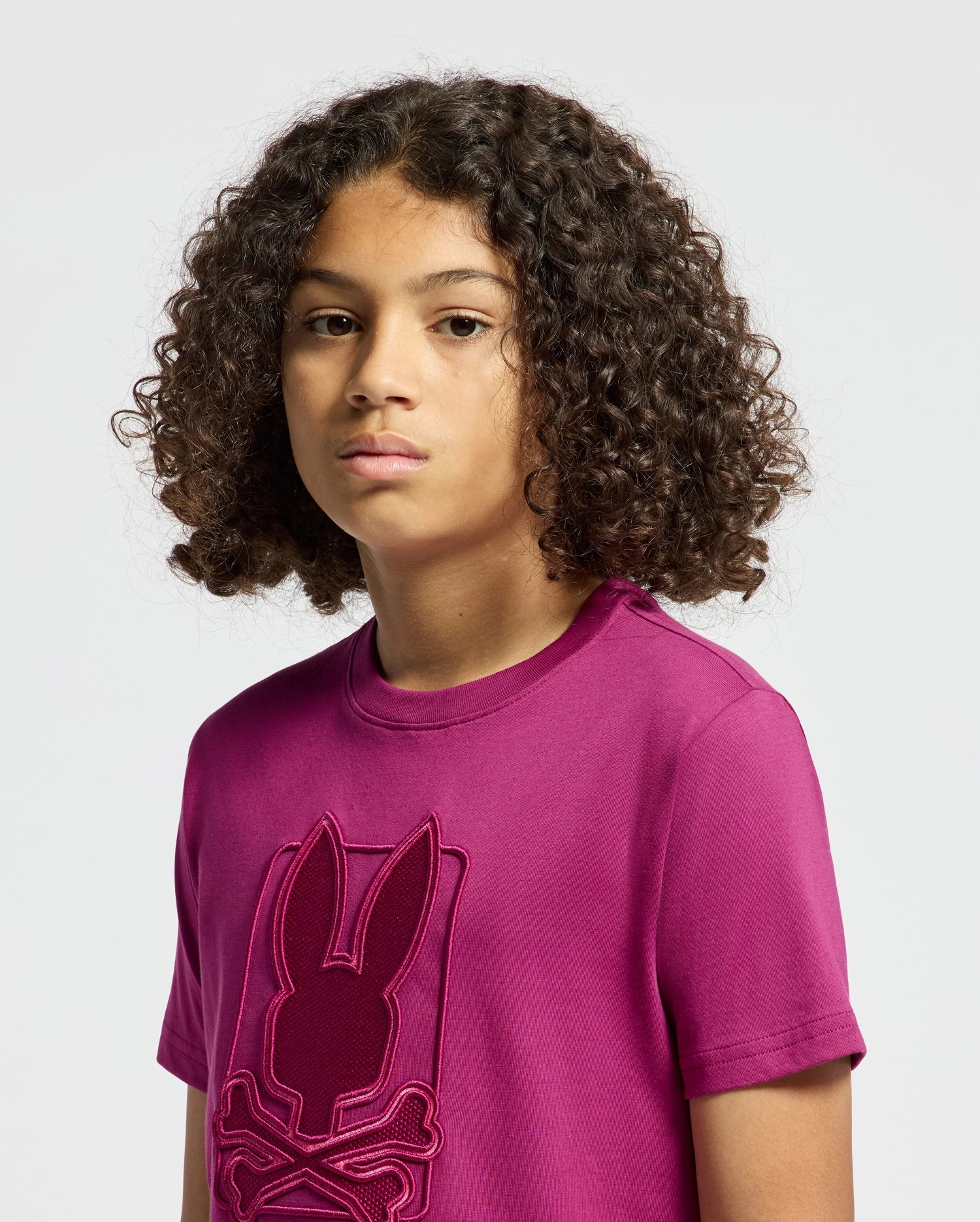 A child with curly hair is wearing a magenta Kids Pierce Graphic Tee by Psycho Bunny, featuring a stylish embroidered bunny graphic. The background is plain and light-colored, complementing the child's neutral expression.