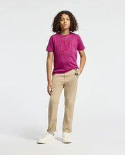A young individual with curly hair exudes confidence in a KIDS PIERCE GRAPHIC TEE - B0U115E200 by Psycho Bunny, featuring a playful bunny design. They complement it with beige pants adorned with an embroidered small character design and finish the outfit with white sneakers. The backdrop is simple and light-colored.
