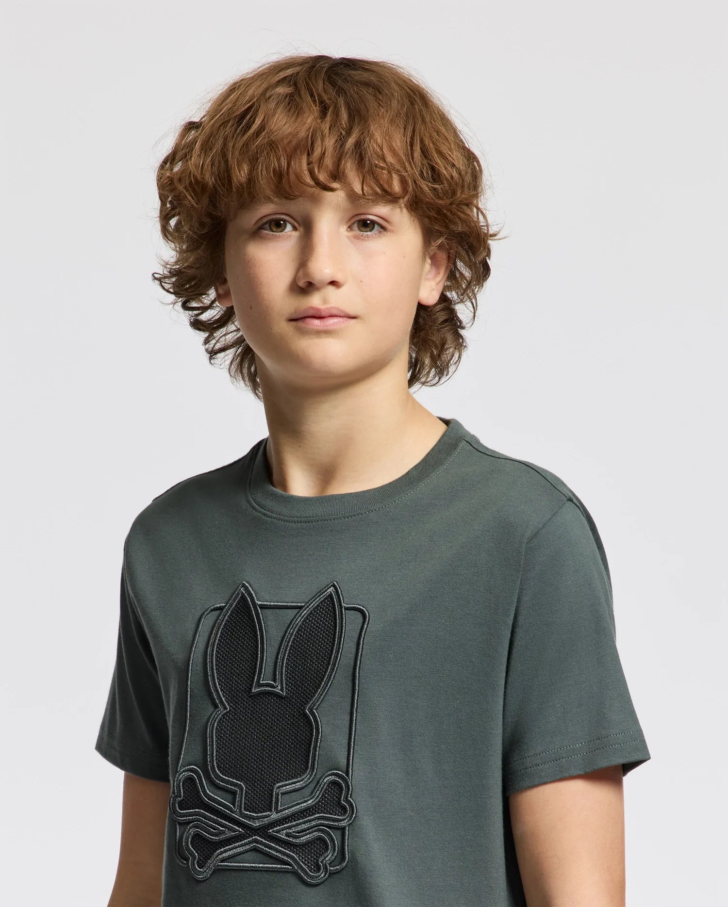 A young person with curly hair is wearing a dark green KIDS PIERCE GRAPHIC TEE - B0U115E200 by Psycho Bunny, which features an embroidered design of a rabbit head and crossed bones. The background is a plain light gray.