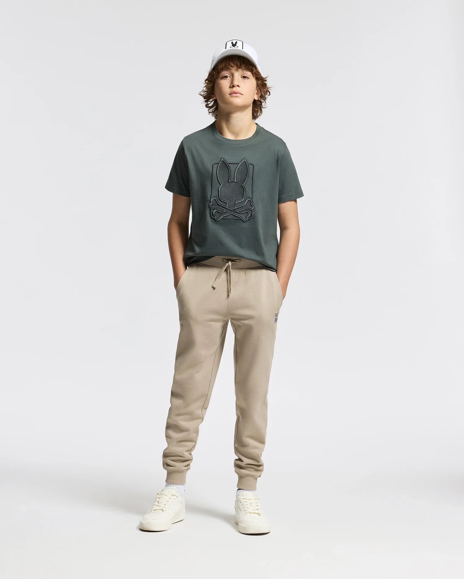 A person wearing a white cap, the Psycho Bunny KIDS PIERCE GRAPHIC TEE in dark green, beige jogger pants, and white sneakers stands against a plain white background. Their hands are in their pockets and they have a relaxed posture.