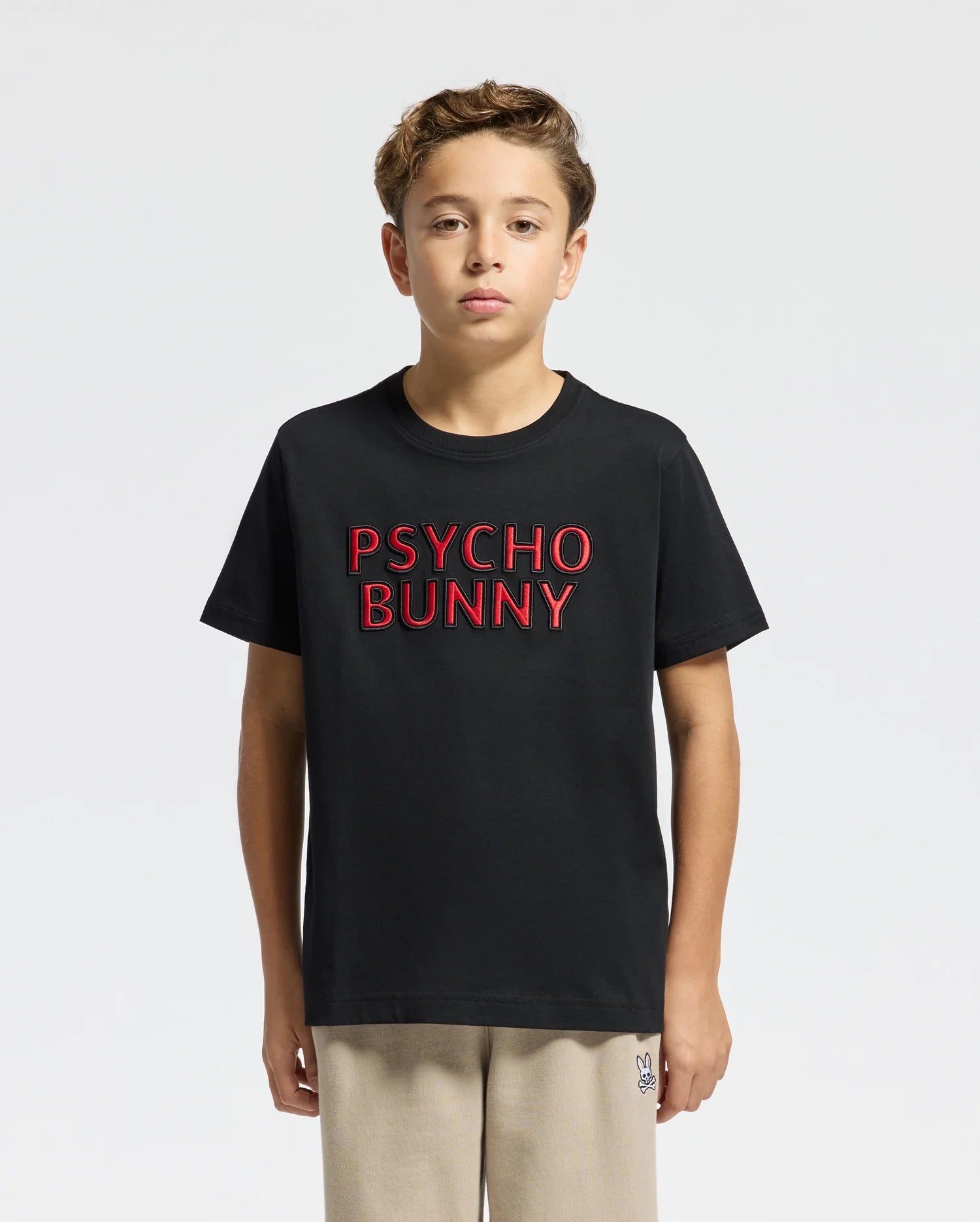 A young person with curly hair sports the KIDS PIERCE EMBROIDERED GRAPHIC TEE - B0U116E200 in black, featuring "Psycho Bunny" in red text, along with beige pants. Made from soft Pima cotton, this tee complements their relaxed style against a plain light background.