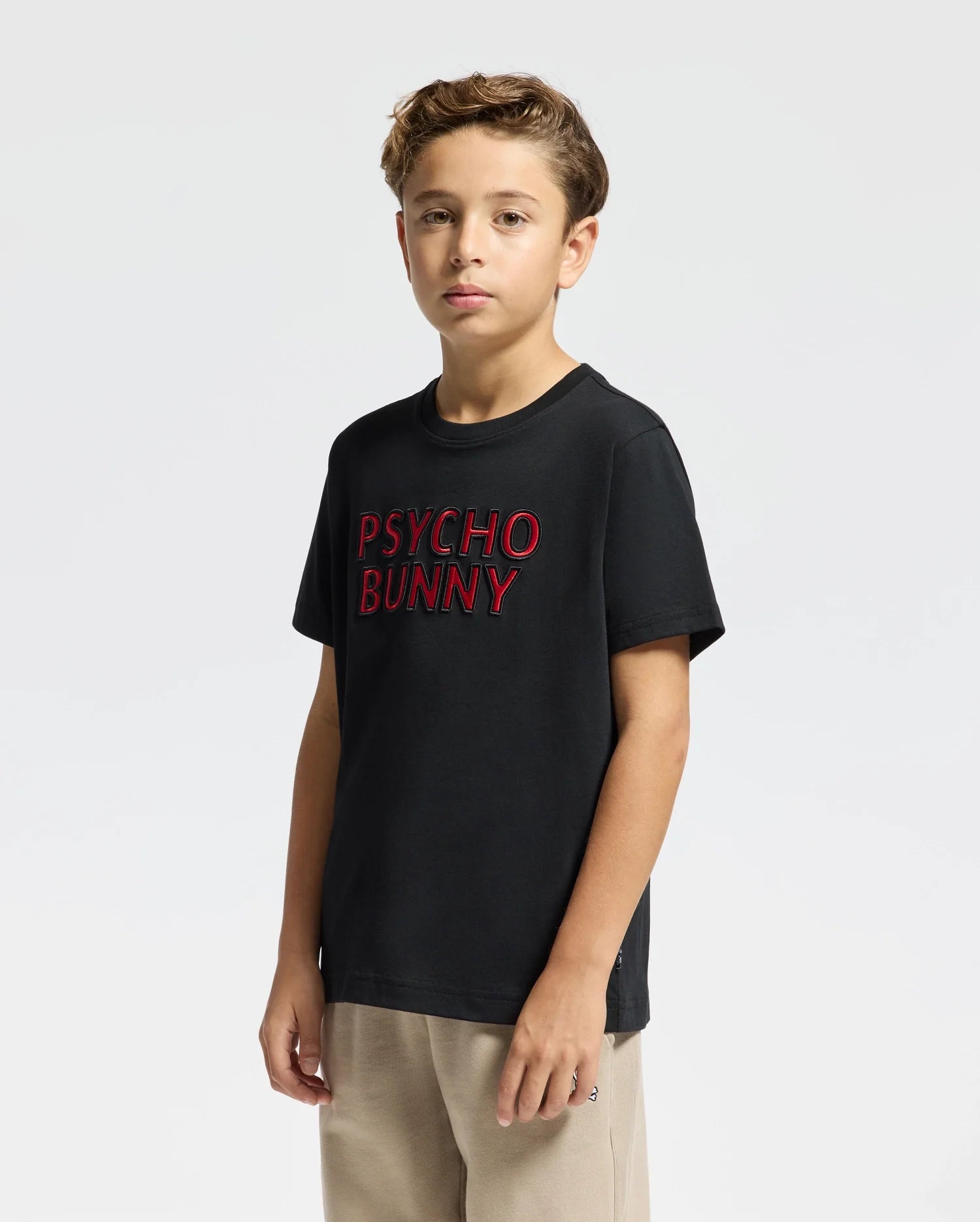 A young person with short brown hair stands against a plain background, wearing the KIDS PIERCE EMBROIDERED GRAPHIC TEE by Psycho Bunny, featuring bold red "PSYCHO BUNNY" text. The look is completed with comfy beige pants.