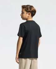 A young boy with short, light brown hair stands looking left in a stylish Psycho Bunny KIDS PIERCE EMBROIDERED GRAPHIC TEE (B0U116E200) made of soft Pima cotton and light-colored pants, set against a plain white background.