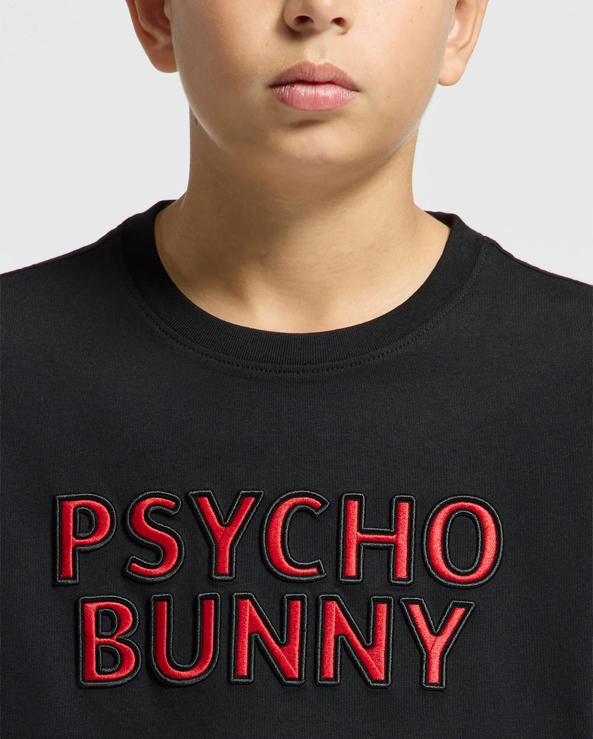 A child wears the KIDS PIERCE EMBROIDERED GRAPHIC TEE by Psycho Bunny, featuring bold red "PSYCHO BUNNY" letters on a black background. Made from soft Pima cotton, it stands out against the plain gray backdrop.