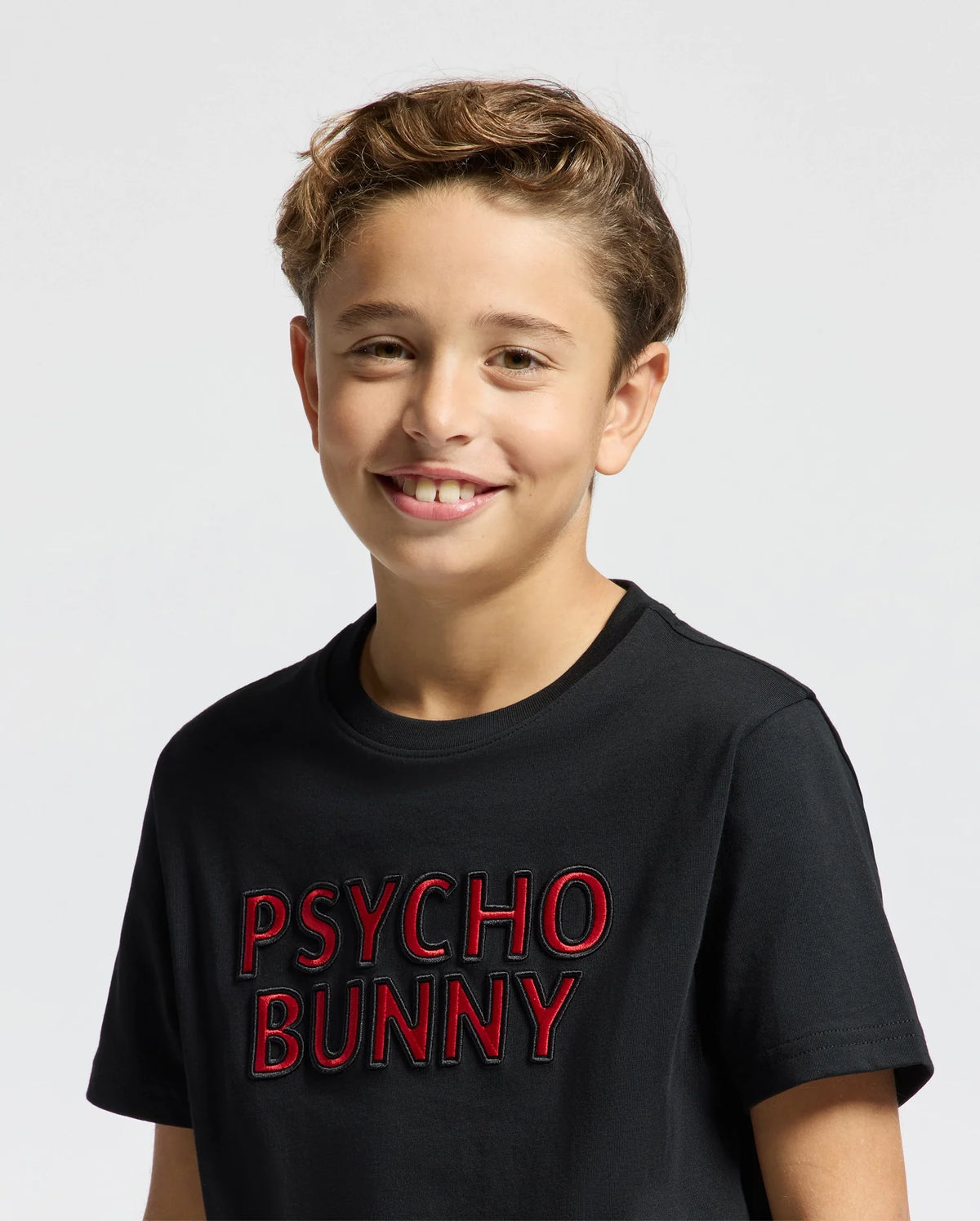 A young person smiles in a black KIDS PIERCE EMBROIDERED GRAPHIC TEE by Psycho Bunny, featuring bold red branding and made from soft pima cotton, against a neutral background.