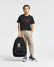 A young boy stands against a plain backdrop, wearing a Psycho Bunny KIDS PIERCE EMBROIDERED GRAPHIC TEE in black with red text made from soft Pima Cotton, beige pants, and black high-tops. He holds a black backpack featuring a skull and bunny logo.
