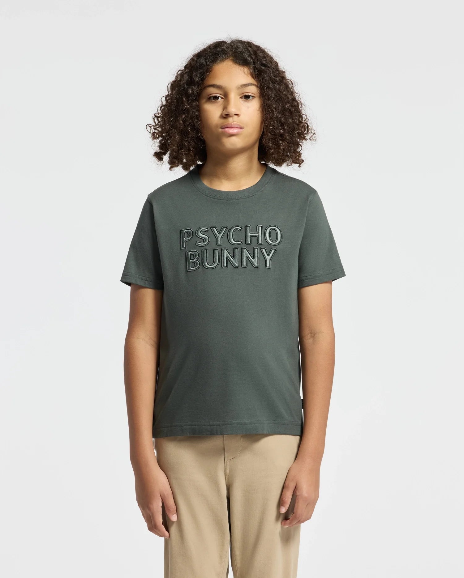 A young individual with curly hair sports a KIDS PIERCE EMBROIDERED GRAPHIC TEE by Psycho Bunny in dark green. Against a plain white background, they effortlessly complement this casual look with beige pants.