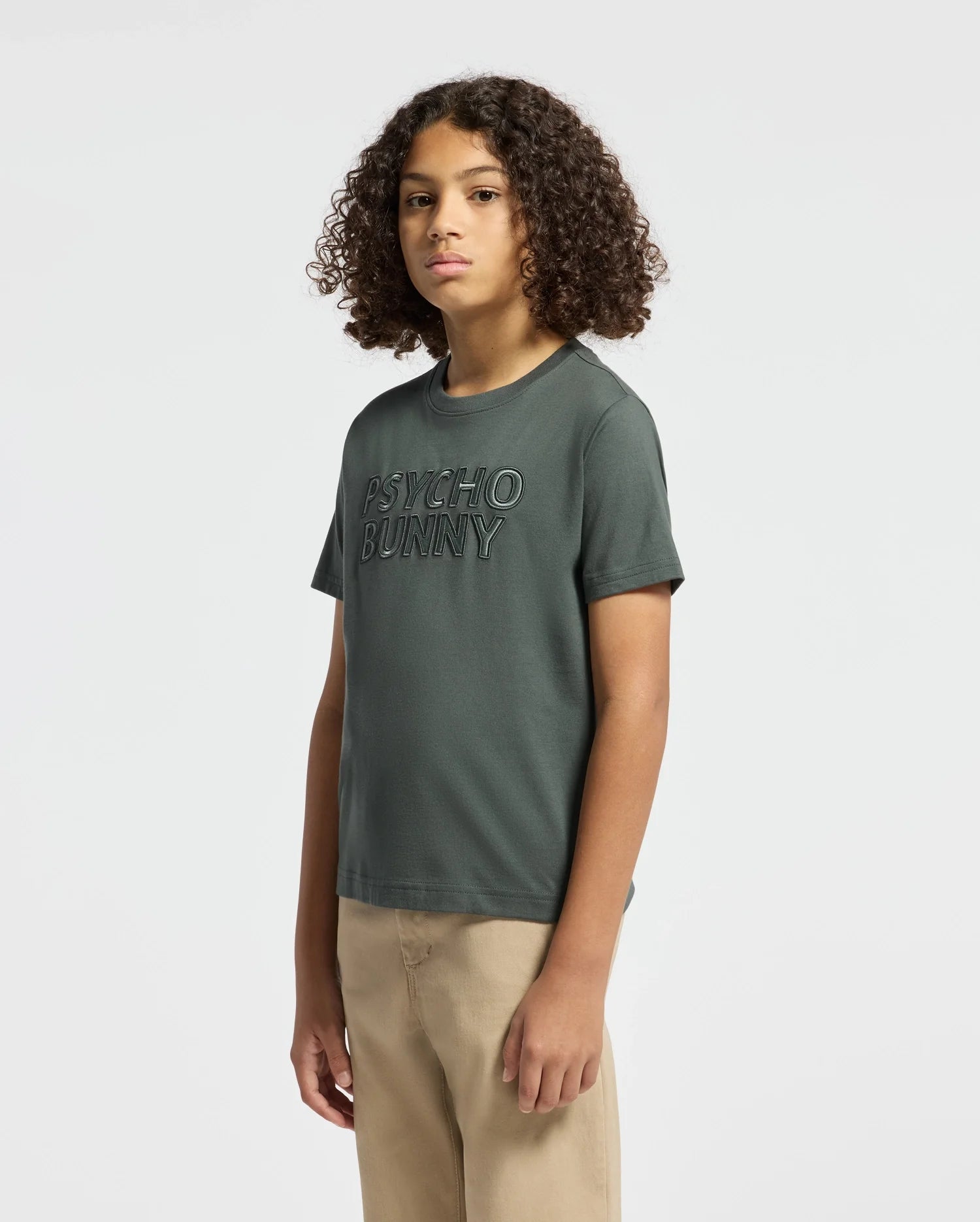 A person with curly hair is wearing a dark green KIDS PIERCE EMBROIDERED GRAPHIC TEE - B0U116E200 by Psycho Bunny, paired with beige pants, standing against a plain white background.