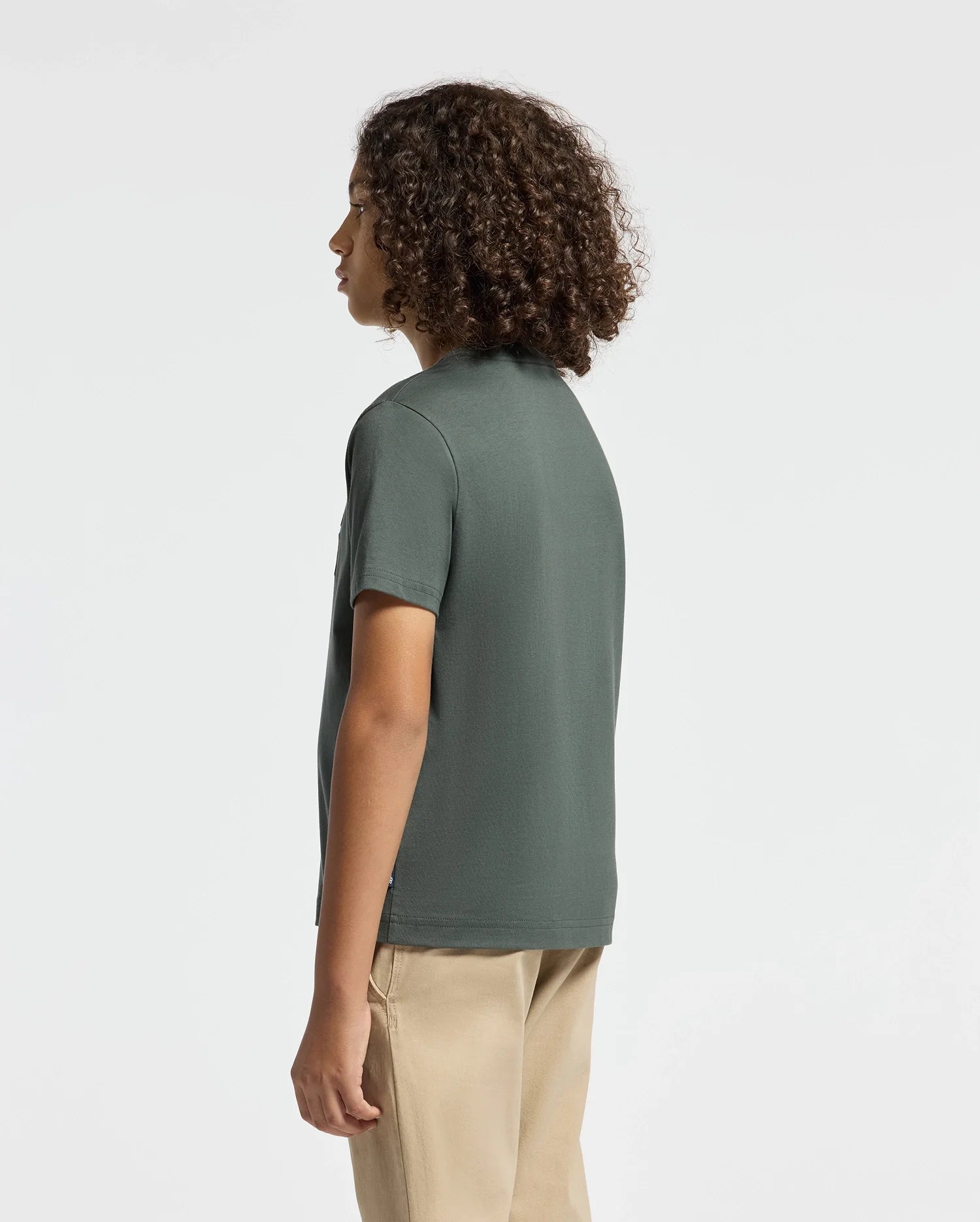 A person with curly hair stands wearing a dark green KIDS PIERCE EMBROIDERED GRAPHIC TEE - B0U116E200 by Psycho Bunny and beige pants, facing slightly to the left in a minimalistic setting.