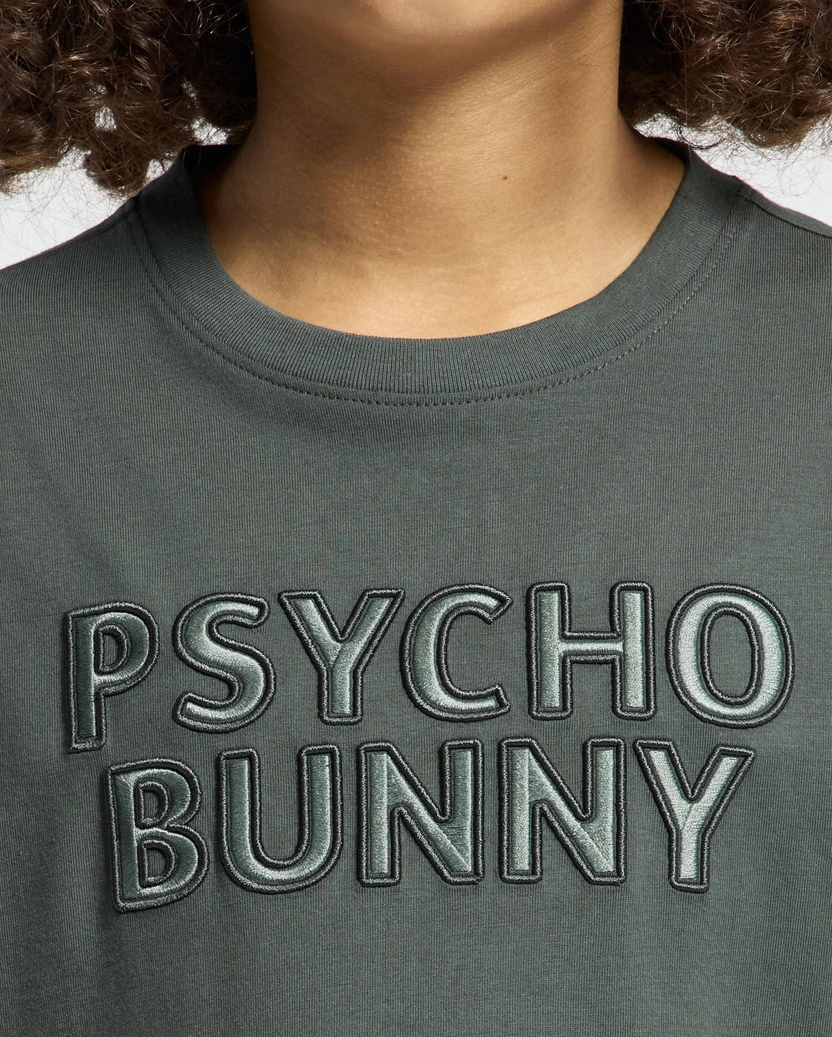 A close-up of a person wearing the KIDS PIERCE EMBROIDERED GRAPHIC TEE - B0U116E200 from Psycho Bunny, crafted from premium Pima cotton in dark green, featuring "PSYCHO BUNNY" embroidered in bold, textured letters on the front. The image highlights the upper section of this stylish T-shirt and its striking text.