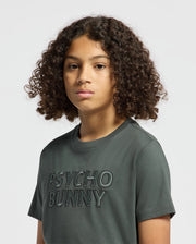 A person with curly hair is wearing a dark green kids' tee, featuring the embroidered text "Psycho Bunny." The shirt is made of a soft fabric that suggests pima cotton, set against a plain light gray background.