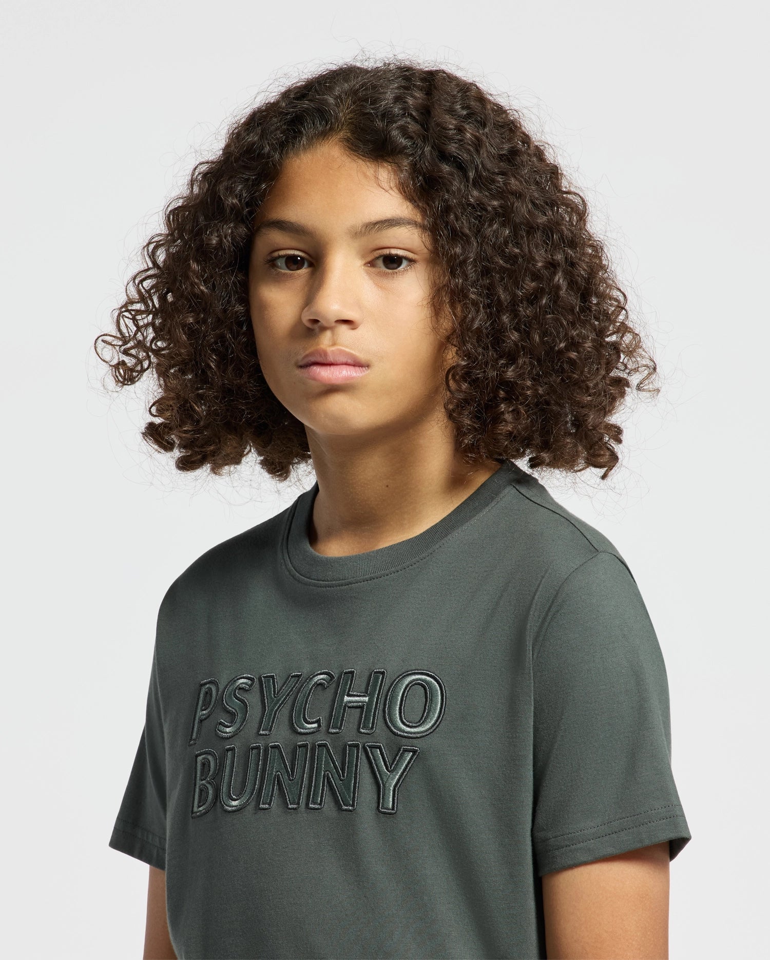 A person with curly hair is wearing a dark green kids' tee, featuring the embroidered text "Psycho Bunny." The shirt is made of a soft fabric that suggests pima cotton, set against a plain light gray background.