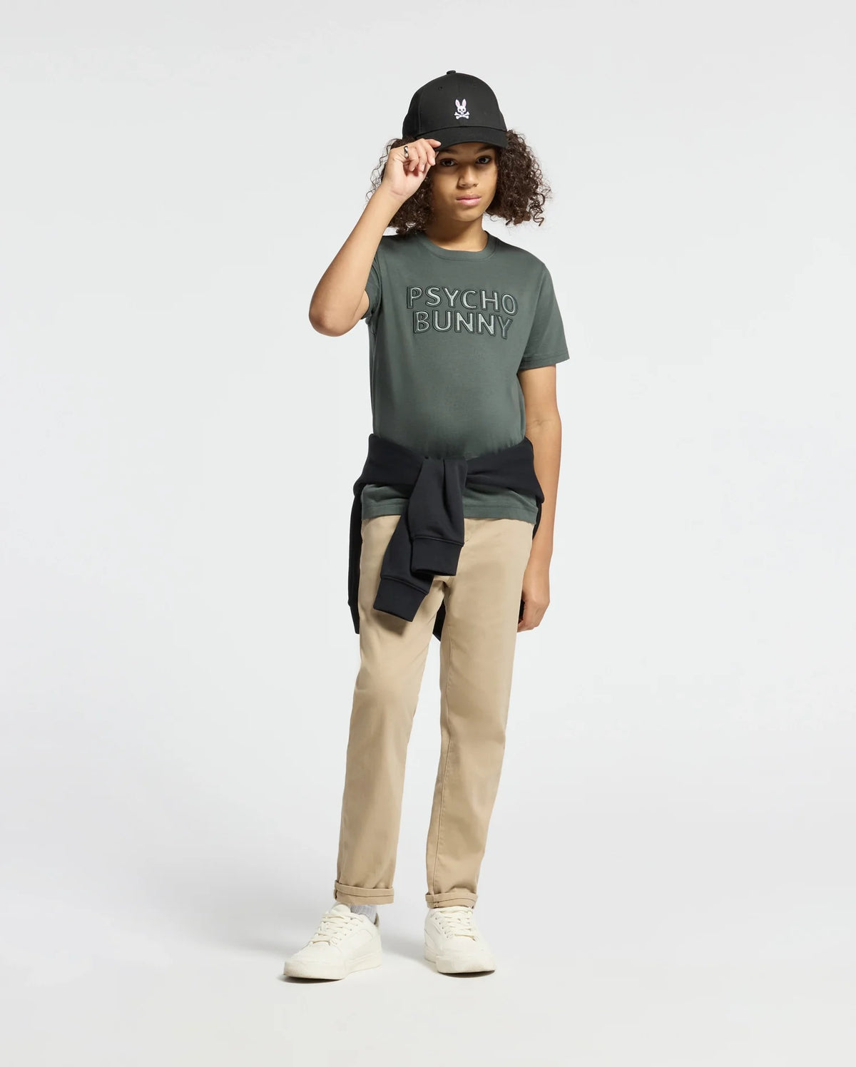 A person wearing a green "KIDS PIERCE EMBROIDERED GRAPHIC TEE - B0U116E200" by Psycho Bunny, paired with khaki pants and white sneakers, touches the brim of a black cap. A black sweatshirt is tied around their waist as they stand against a plain white background, highlighting the smooth texture of pima cotton.