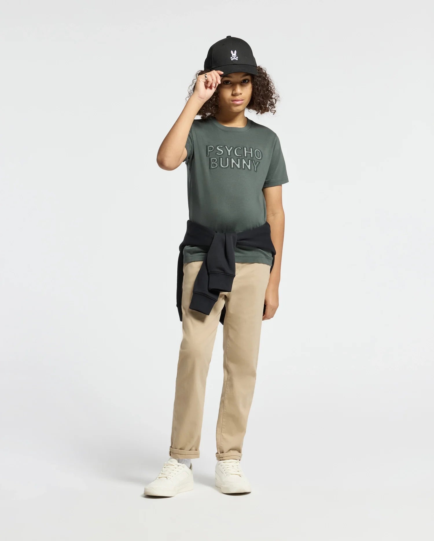 A person wearing a green "KIDS PIERCE EMBROIDERED GRAPHIC TEE - B0U116E200" by Psycho Bunny, paired with khaki pants and white sneakers, touches the brim of a black cap. A black sweatshirt is tied around their waist as they stand against a plain white background, highlighting the smooth texture of pima cotton.