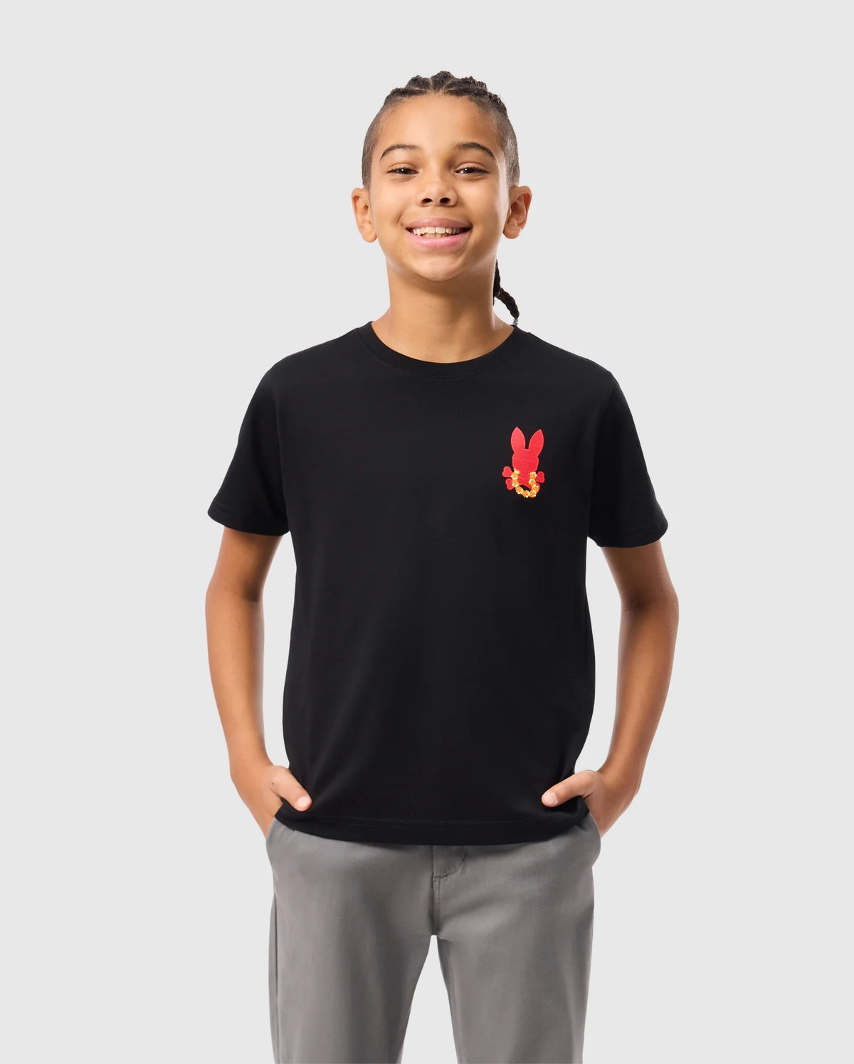 A child with braided hair is wearing the KIDS HAWAII CITY TEE from Psycho Bunny, a black Pima cotton t-shirt with a red bunny graphic, paired with gray pants. They stand hands in pockets against a plain white background.