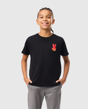 A child with braided hair is wearing the KIDS HAWAII CITY TEE from Psycho Bunny, a black Pima cotton t-shirt with a red bunny graphic, paired with gray pants. They stand hands in pockets against a plain white background.