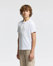 A young person with curly hair stands against a plain background, wearing the KIDS DOTSERO PRINTED GRAPHIC TEE by Psycho Bunny, crafted from Peruvian Pima Cotton and featuring a puff print graphic. They pair it with beige pants and gaze straight at the camera with a neutral expression.
