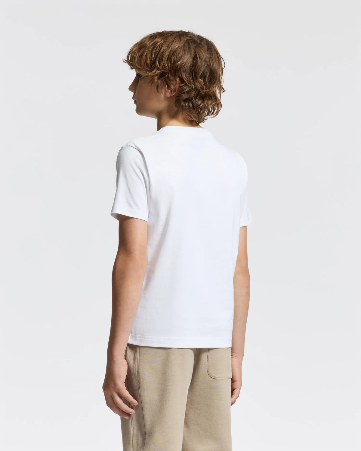 A young individual with short, wavy hair is facing away, dressed in a Psycho Bunny KIDS DOTSERO PRINTED GRAPHIC TEE - B0U197E200 and light-colored pants. The background is neutral and minimalistic.