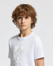 A young boy with curly brown hair wears the Psycho Bunny KIDS DOTSERO PRINTED GRAPHIC TEE in white, featuring a puff print graphic design on the front. Made from soft Peruvian Pima Cotton, he gazes at the camera with a neutral expression against a plain light background.