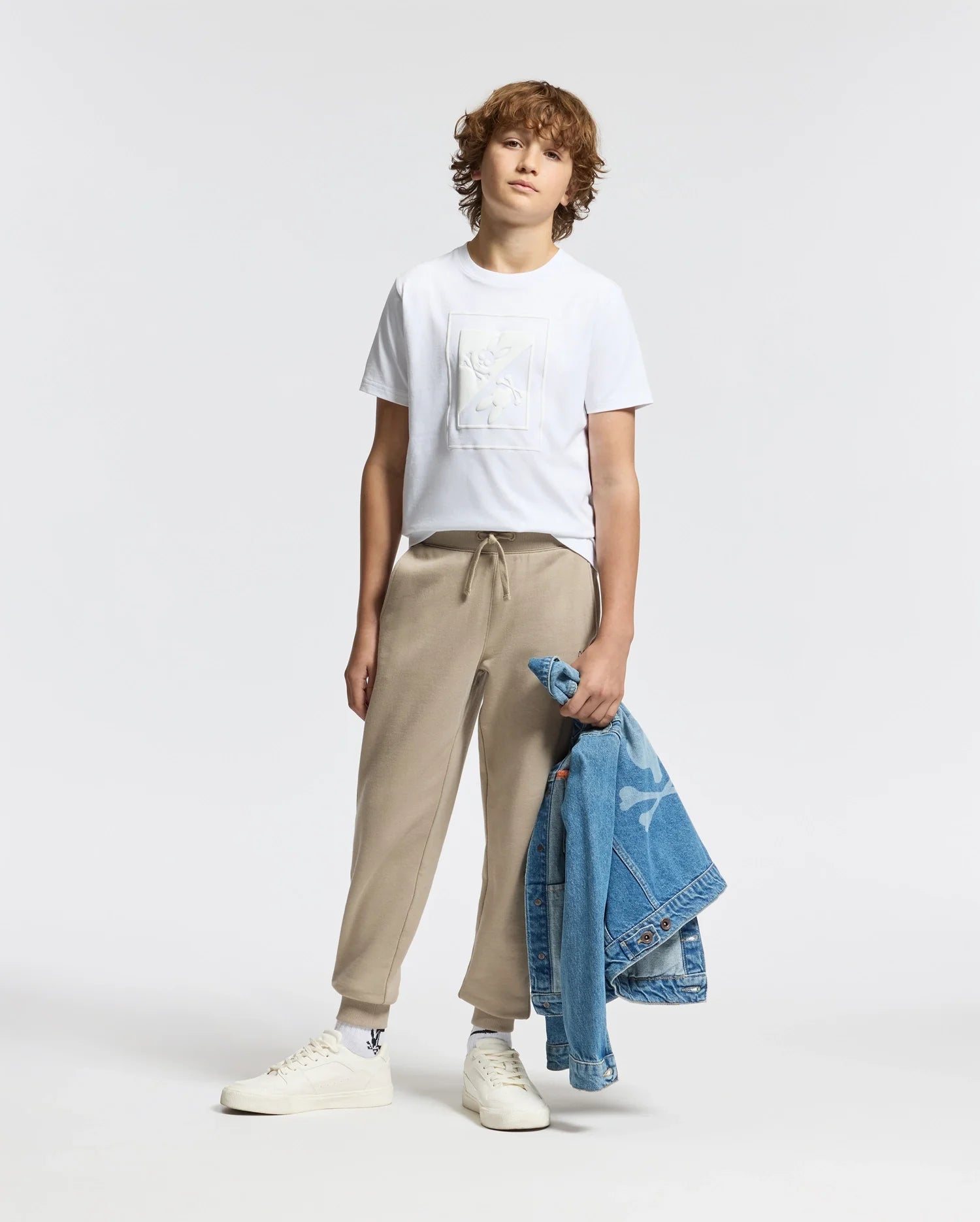 A young person with curly hair stands against a plain background, wearing a white KIDS DOTSERO PRINTED GRAPHIC TEE by Psycho Bunny and beige joggers. They hold a blue denim jacket in one hand and wear white sneakers.