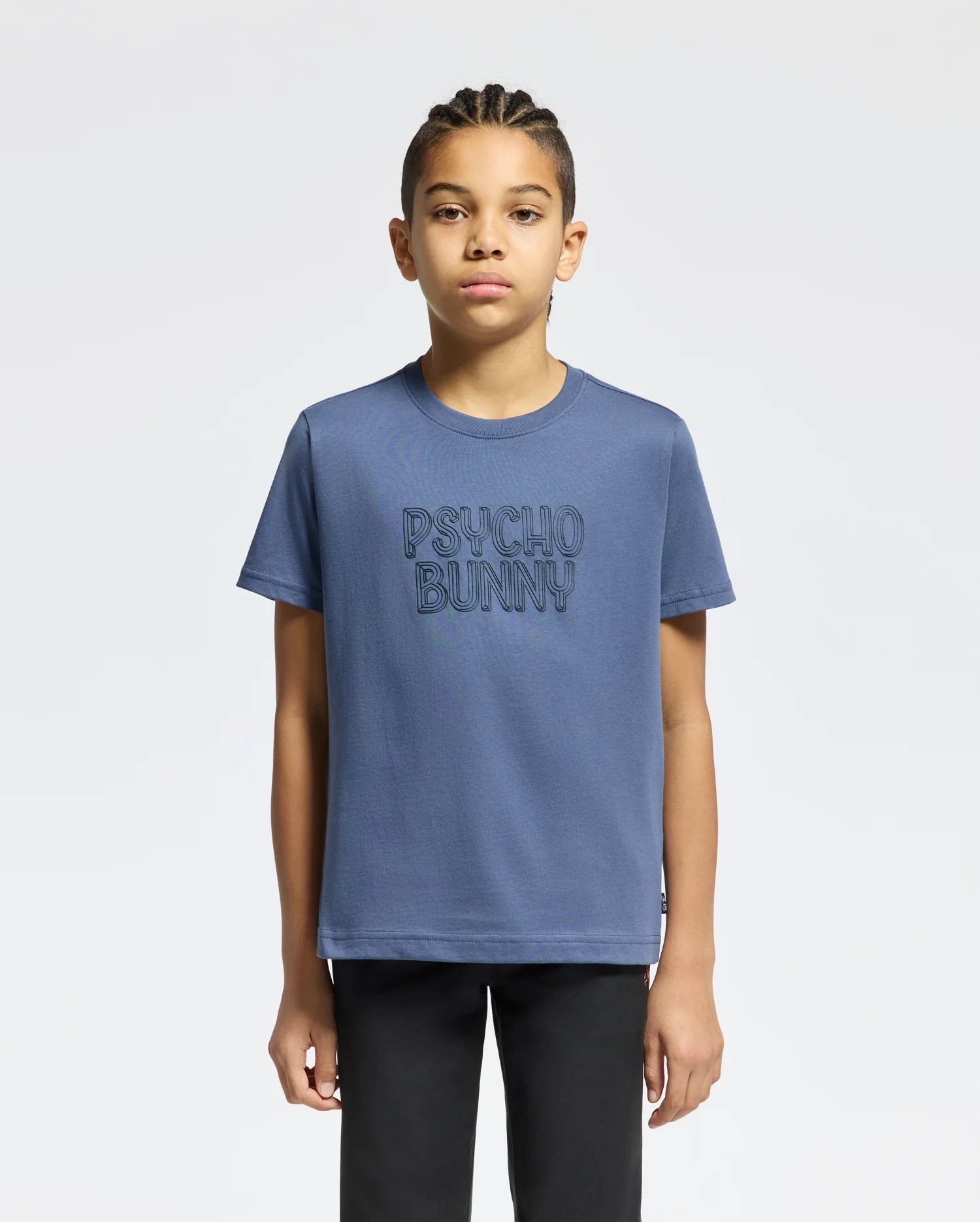 A young individual with braided hair is sporting the KIDS MONARC WORDMARK GRAPHIC TEE - B0U210E200 by Psycho Bunny, made from lightweight and durable Pima cotton, featuring the "PSYCHO BUNNY" text on the front. They stand confidently against a plain white background.