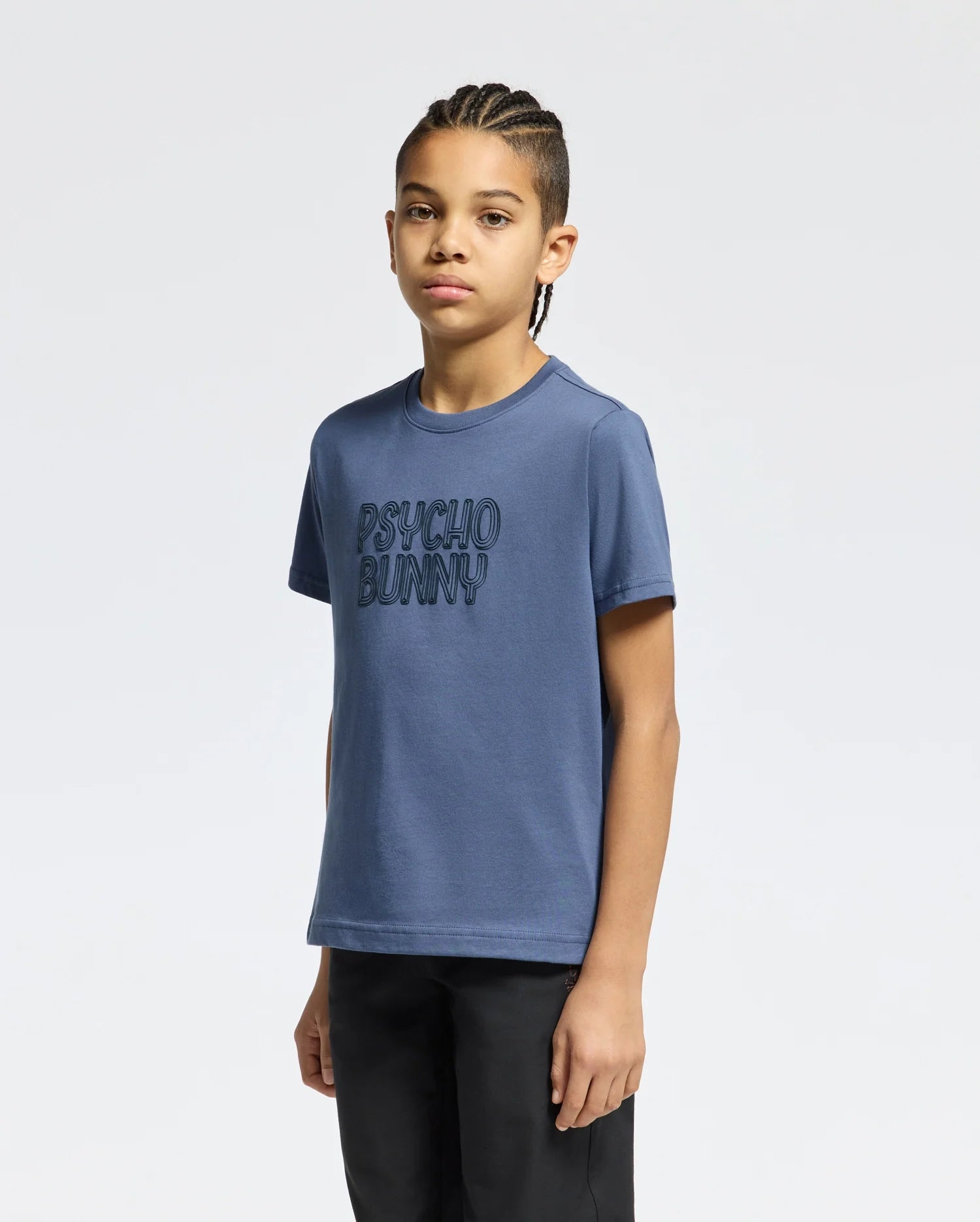 A boy with braided hair is wearing the Kids Monarc Wordmark Graphic Tee - B0U210E200, a lightweight and durable blue T-shirt crafted from soft Pima cotton, showcasing the "Psycho Bunny" design on the front. He stands against a plain white background with a neutral expression.