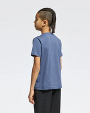 A child with braided hair stands sideways, dressed in a KIDS MONARC WORDMARK GRAPHIC TEE - B0U210E200 made from lightweight, durable Pima cotton by Psycho Bunny and paired with black pants, all set against a plain white background.