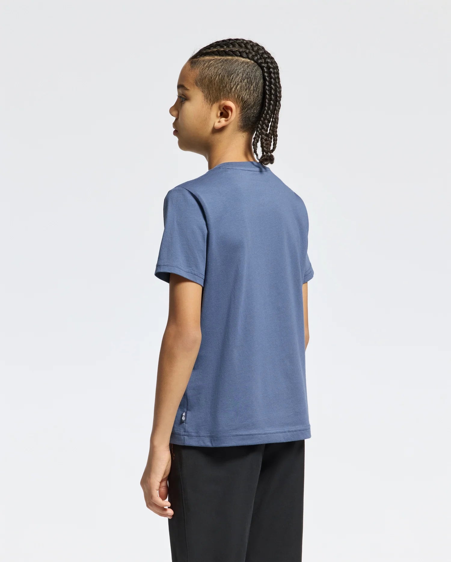 A child with braided hair stands sideways, dressed in a KIDS MONARC WORDMARK GRAPHIC TEE - B0U210E200 made from lightweight, durable Pima cotton by Psycho Bunny and paired with black pants, all set against a plain white background.
