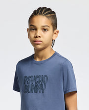 A young person with braided hair wears the Kids Monarc Wordmark Graphic Tee in blue, crafted from lightweight and durable Pima cotton by Psycho Bunny, standing against a plain white background.