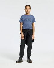 A young person with braided hair stands against a plain background wearing a blue KIDS MONARC WORDMARK GRAPHIC TEE from Psycho Bunny, crafted from lightweight, durable Pima cotton. They are also dressed in black pants and black shoes, with a black jacket tied around their waist.