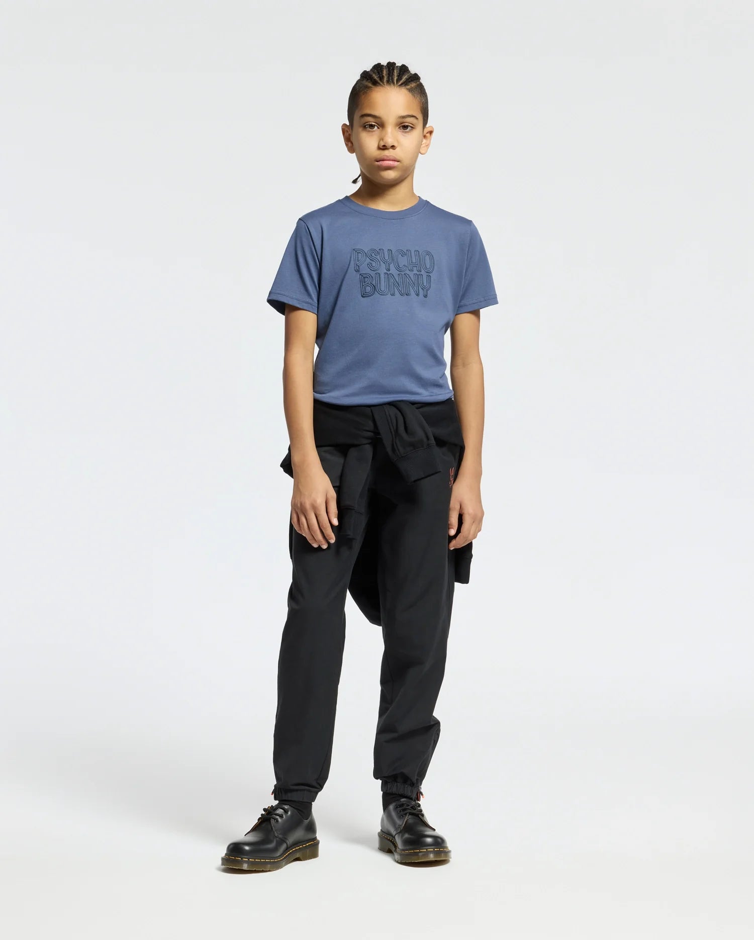 A young person with braided hair stands against a plain background wearing a blue KIDS MONARC WORDMARK GRAPHIC TEE from Psycho Bunny, crafted from lightweight, durable Pima cotton. They are also dressed in black pants and black shoes, with a black jacket tied around their waist.