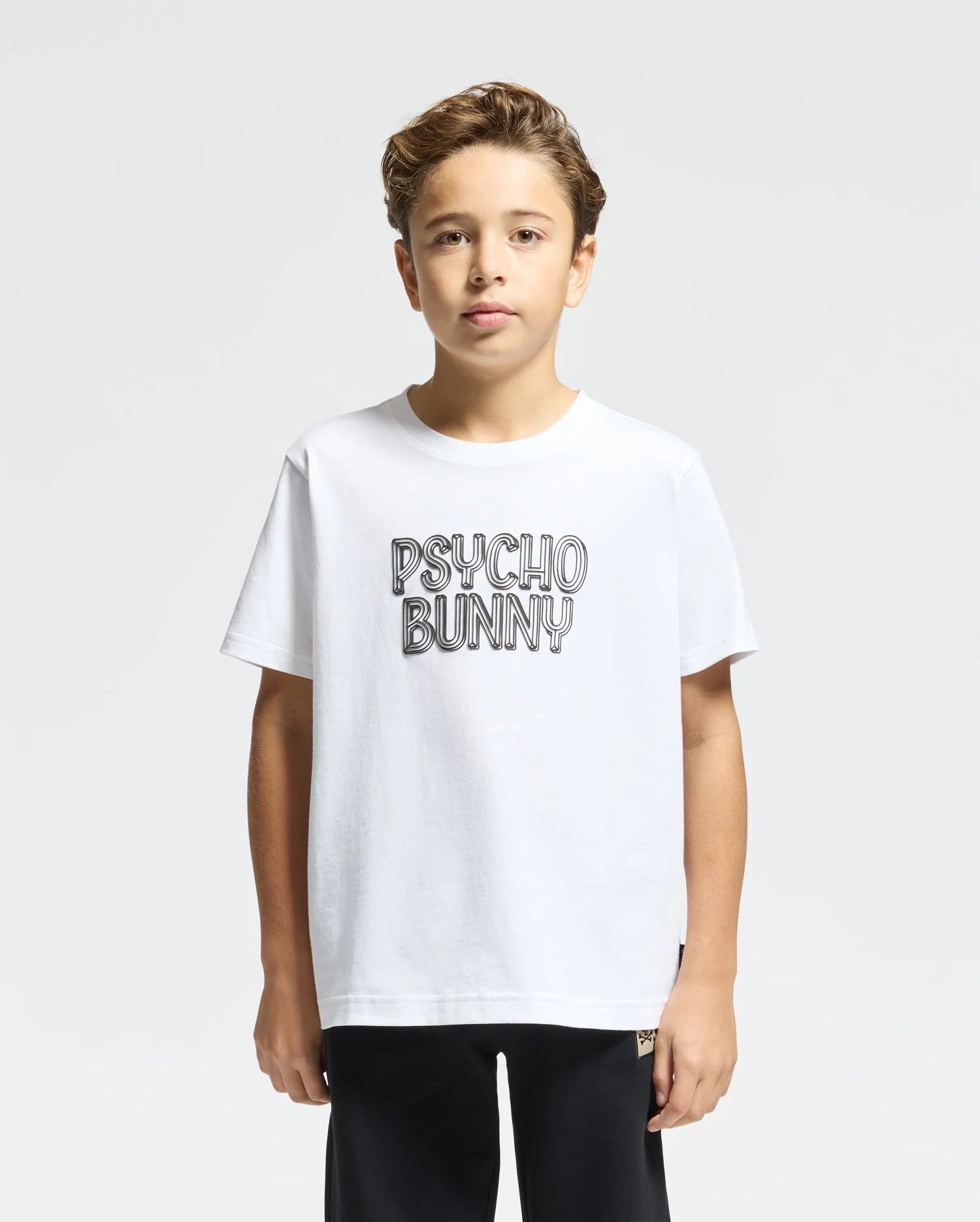 A young individual with short hair is wearing the "KIDS MONARC WORDMARK GRAPHIC TEE - B0U210E200" by Psycho Bunny, crafted from white Pima cotton. They enjoy cocoon comfort as they stand against a plain gray background.