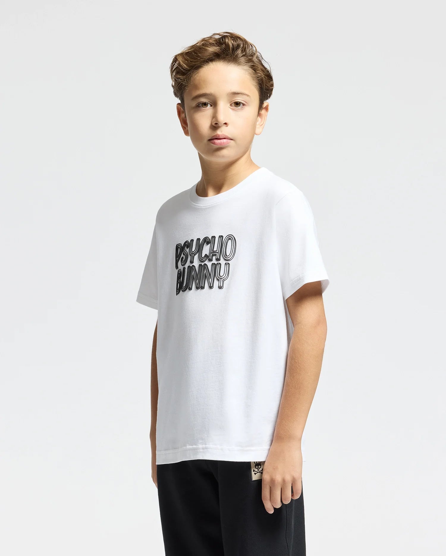 A young boy stands wearing the Kids Monarc Wordmark Graphic Tee by Psycho Bunny, crafted from soft Pima cotton with the bold text "PSYCHO BUNNY" printed on the front. His short, wavy hair complements his dark pants, creating a stylish ensemble against the plain, light-colored background.