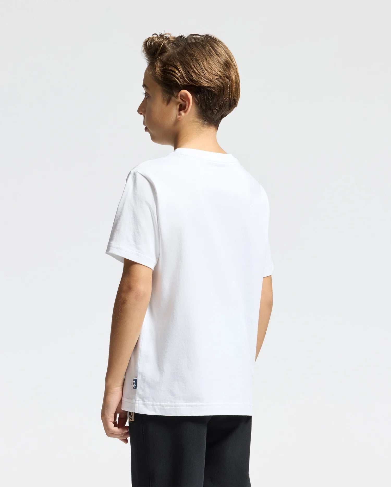 A young boy with light brown hair stands against a plain white background, wearing a Psycho Bunny KIDS MONARC WORDMARK GRAPHIC TEE - B0U210E200 made of soft Pima cotton and dark pants, facing slightly to the side.