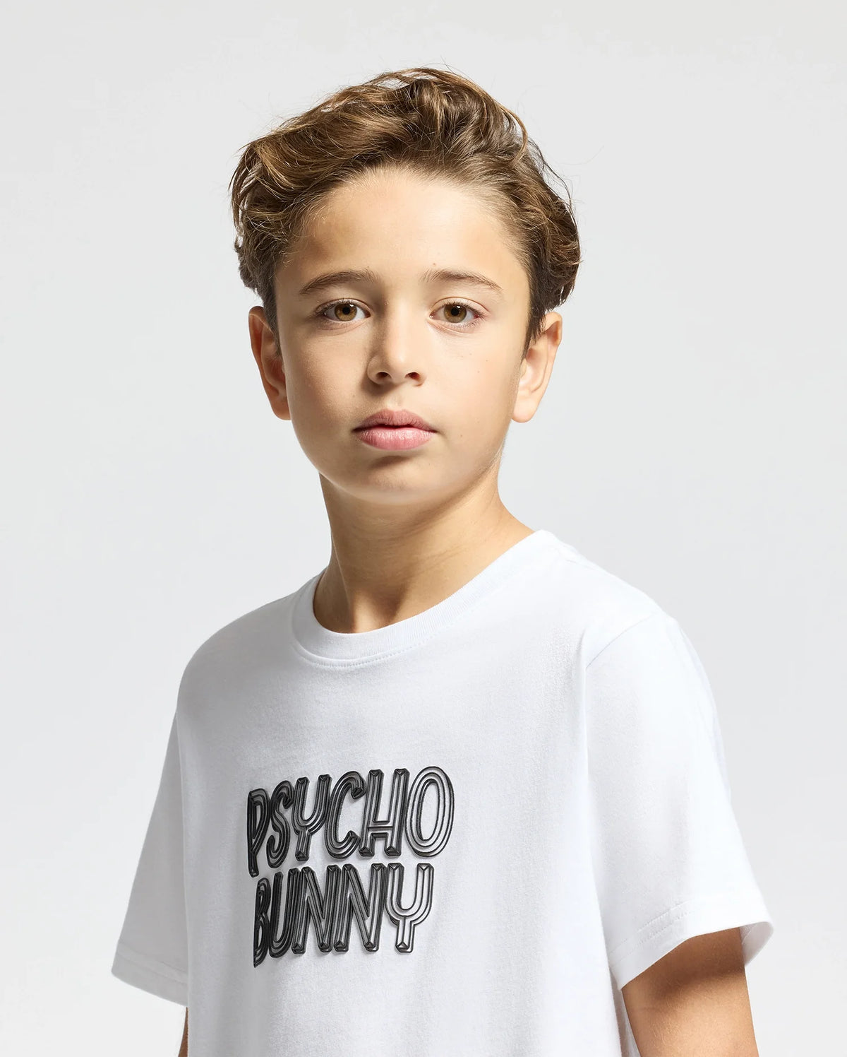 A young boy with short, wavy brown hair is wearing a KIDS MONARC WORDMARK GRAPHIC TEE - B0U210E200 by Psycho Bunny, crafted from soft Pima cotton. The words "Psycho Bunny" are boldly printed on it as he looks directly at the camera against a plain, light gray background.