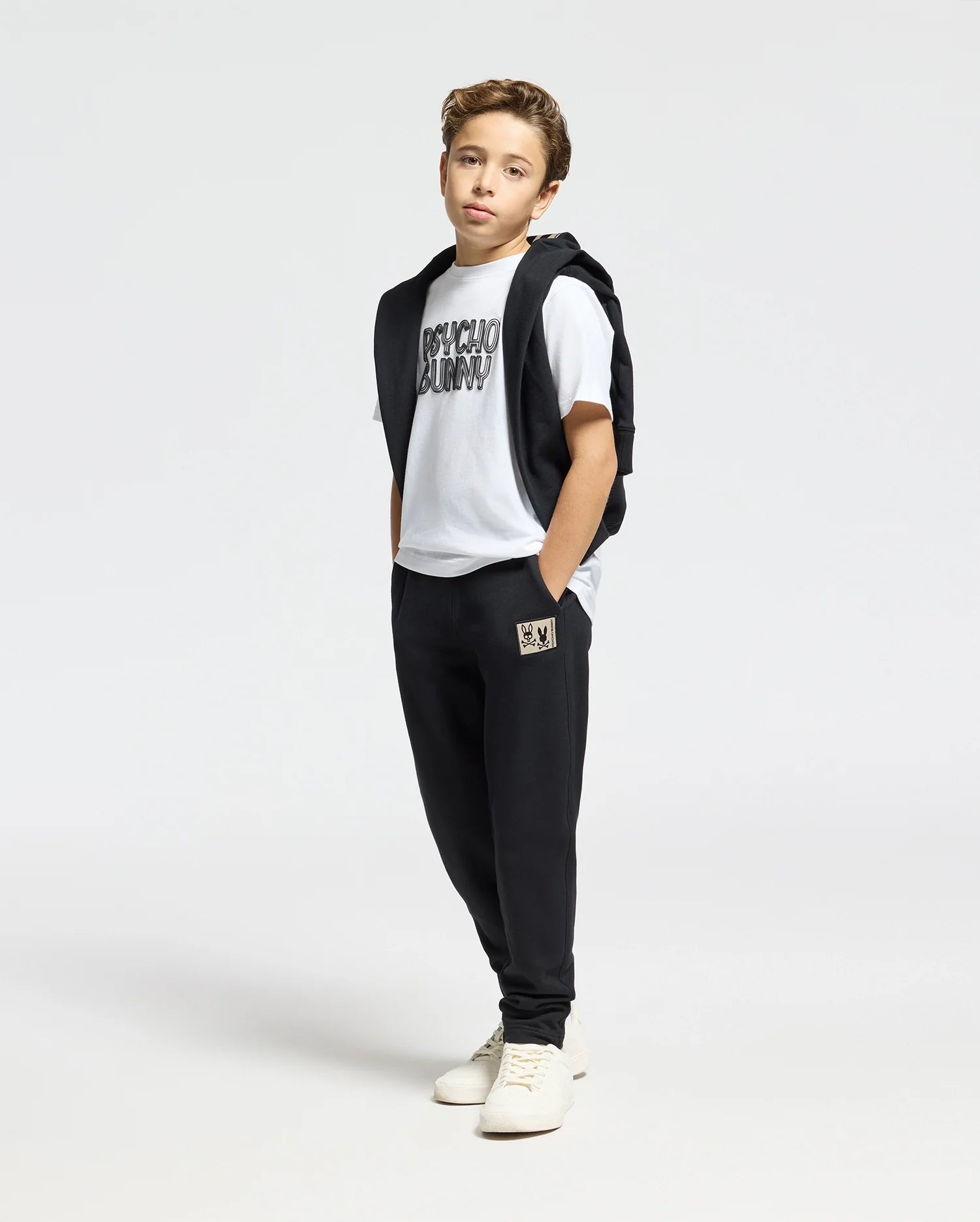 A young boy stands against a plain background wearing a Psycho Bunny KIDS MONARC WORDMARK GRAPHIC TEE crafted from soft Pima cotton, black sweatpants, and a sleeveless black jacket. His hands are in his pockets, and he is comfortably sporting white sneakers.