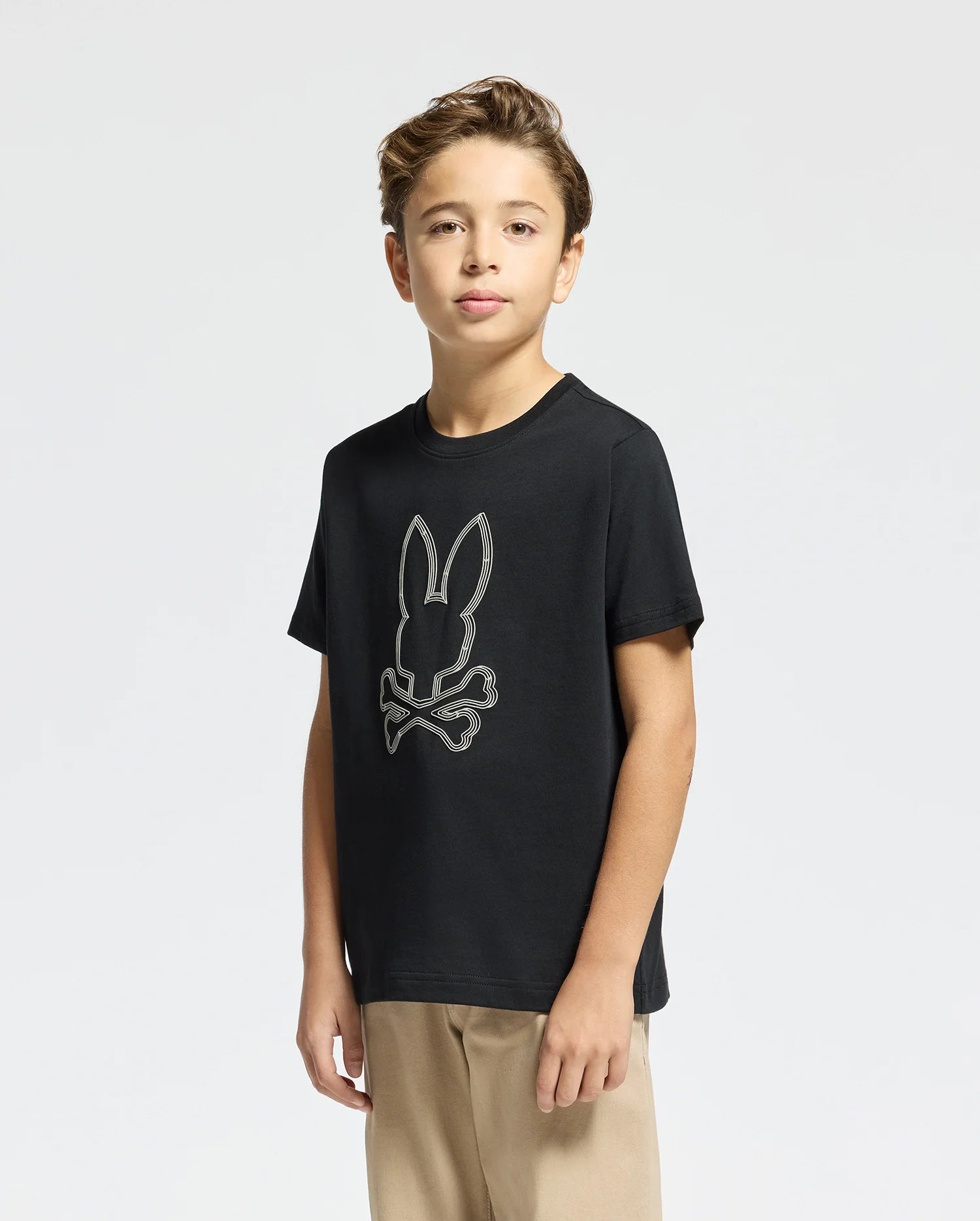 A young person stands confidently wearing the black KIDS MONARC FRONT BUNNY GRAPHIC TEE - B0U211E200 by Psycho Bunny, showcasing a bold bunny and crossbones design. Crafted from soft Pima cotton, the T-shirt complements their beige pants perfectly. Their short brown hair contrasts against a plain white background, embodying a modern Monarc aesthetic.
