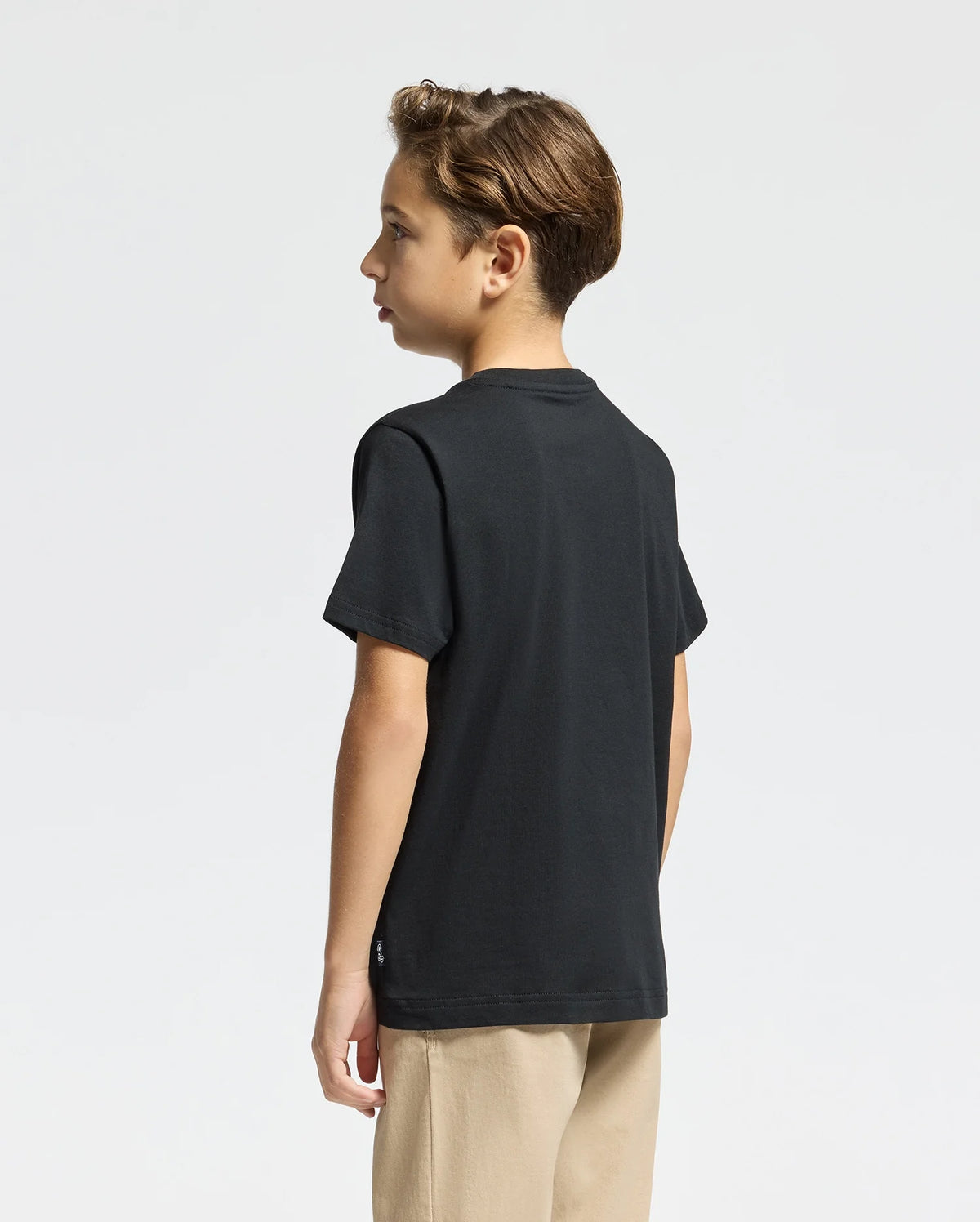 A young boy with short brown hair stands sideways, wearing the KIDS MONARC FRONT BUNNY GRAPHIC TEE - B0U211E200 from Psycho Bunny, crafted from soft Pima cotton, paired with beige pants against a plain white background.