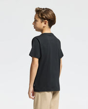 A young boy with short brown hair stands sideways, wearing the KIDS MONARC FRONT BUNNY GRAPHIC TEE - B0U211E200 from Psycho Bunny, crafted from soft Pima cotton, paired with beige pants against a plain white background.