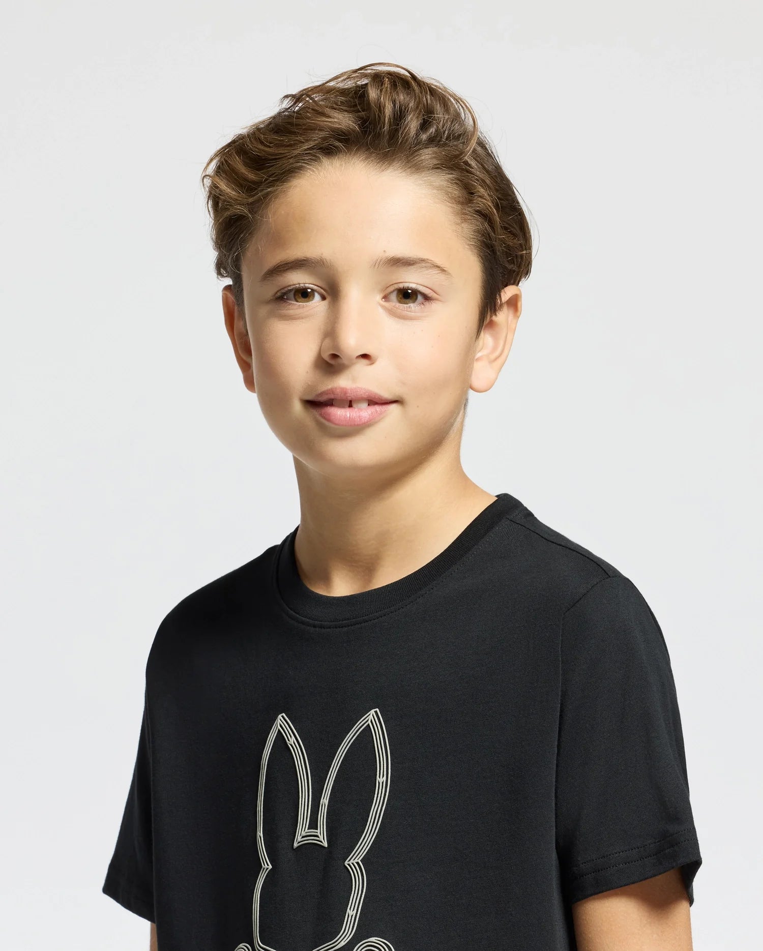 A young boy with short, light brown hair is sporting a black Kids Monarc Front Bunny Graphic Tee, made from soft Pima cotton by Psycho Bunny. The tee's simple bunny design adds a touch of charm as he smiles softly against a plain light gray background.