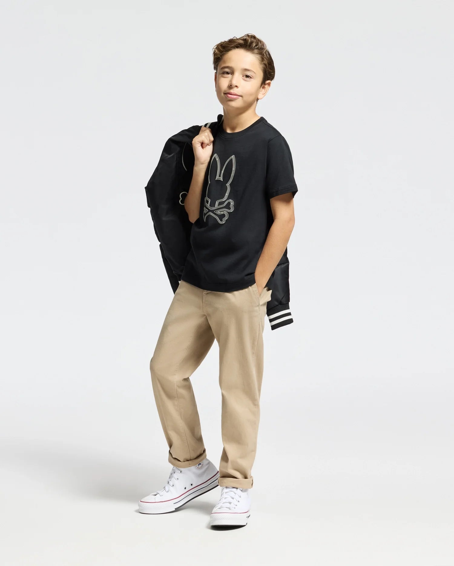 A young person poses confidently, wearing a Psycho Bunny KIDS MONARC FRONT BUNNY GRAPHIC TEE made from soft Pima cotton, tan pants, and white sneakers. They hold a black jacket over their shoulder and stand against a plain background.