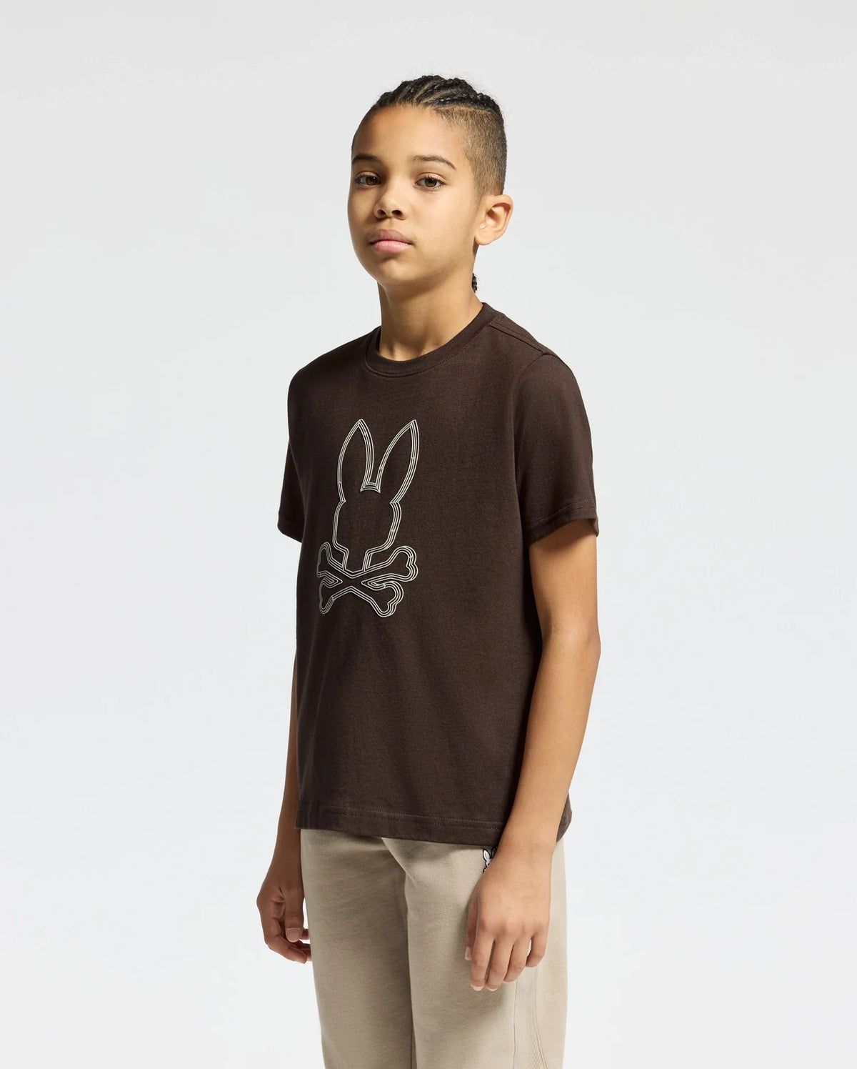 A young person stands confidently in a Psycho Bunny KIDS MONARC FRONT BUNNY GRAPHIC TEE - B0U211E200, crafted from soft Pima Cotton and featuring a bunny skull and crossbones design. They have long braided hair and are dressed in beige pants against a plain white background.