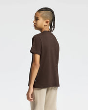A young person with braided hair is standing and looking to the side, wearing the KIDS MONARC FRONT BUNNY GRAPHIC TEE by Psycho Bunny in dark brown. Paired with light-colored pants, they stand against a plain, light-colored backdrop.