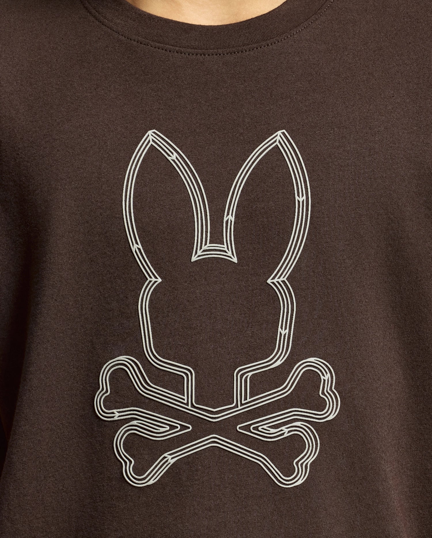 Introducing the KIDS MONARC FRONT BUNNY GRAPHIC TEE - B0U211E200 from Psycho Bunny: a stylish brown shirt crafted from luxurious Pima cotton. It showcases a unique design featuring a stylized outline of a bunny head above crossed bones in crisp white, perfectly capturing the distinctive flair of Monarc.