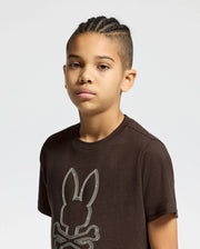 A young person with braided hair is wearing the KIDS MONARC FRONT BUNNY GRAPHIC TEE (B0U211E200) by Psycho Bunny, a brown shirt made of soft Pima Cotton that showcases a stylized bunny and crossbones design. The light-colored background accentuates their casual yet trendy appearance.
