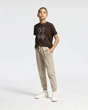 A young person stands confidently with hands in their pockets, wearing a KIDS MONARC FRONT BUNNY GRAPHIC TEE by Psycho Bunny and beige jogger pants. Their ensemble is completed with white sneakers and a necklace, set against a simple light background.