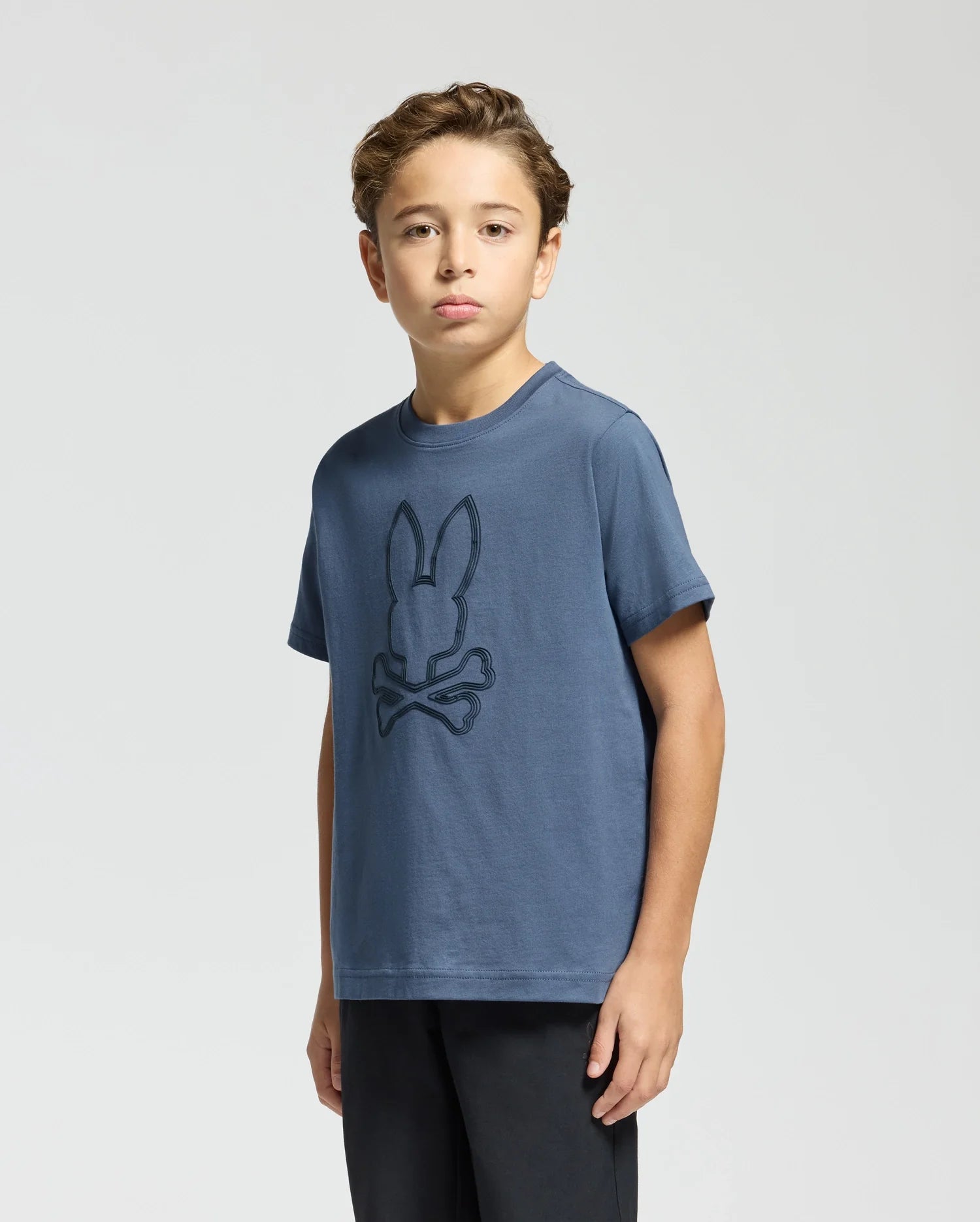 The boy is wearing a Psycho Bunny Kids Monarc Front Bunny Graphic Tee, crafted from soft Pima cotton and displaying a rabbit and crossbones design. He stands against a plain light background, looking directly at the camera.