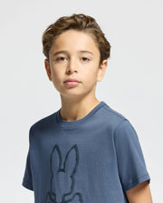 A young boy with light brown hair stands against a light gray background, wearing the Psycho Bunny Kids Monarc Front Bunny Graphic Tee (B0U211E200) in blue, subtly flaunting the iconic bunny design. His expression is neutral.