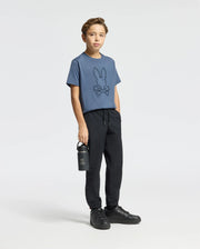 A boy stands against a plain background wearing the Psycho Bunny KIDS MONARC FRONT BUNNY GRAPHIC TEE in blue, crafted from soft Pima cotton and featuring the Monarc Front Bunny logo. He's styled with black jogger pants and sneakers, holding a matching black water bottle with the logo while his other hand rests casually in his pocket.