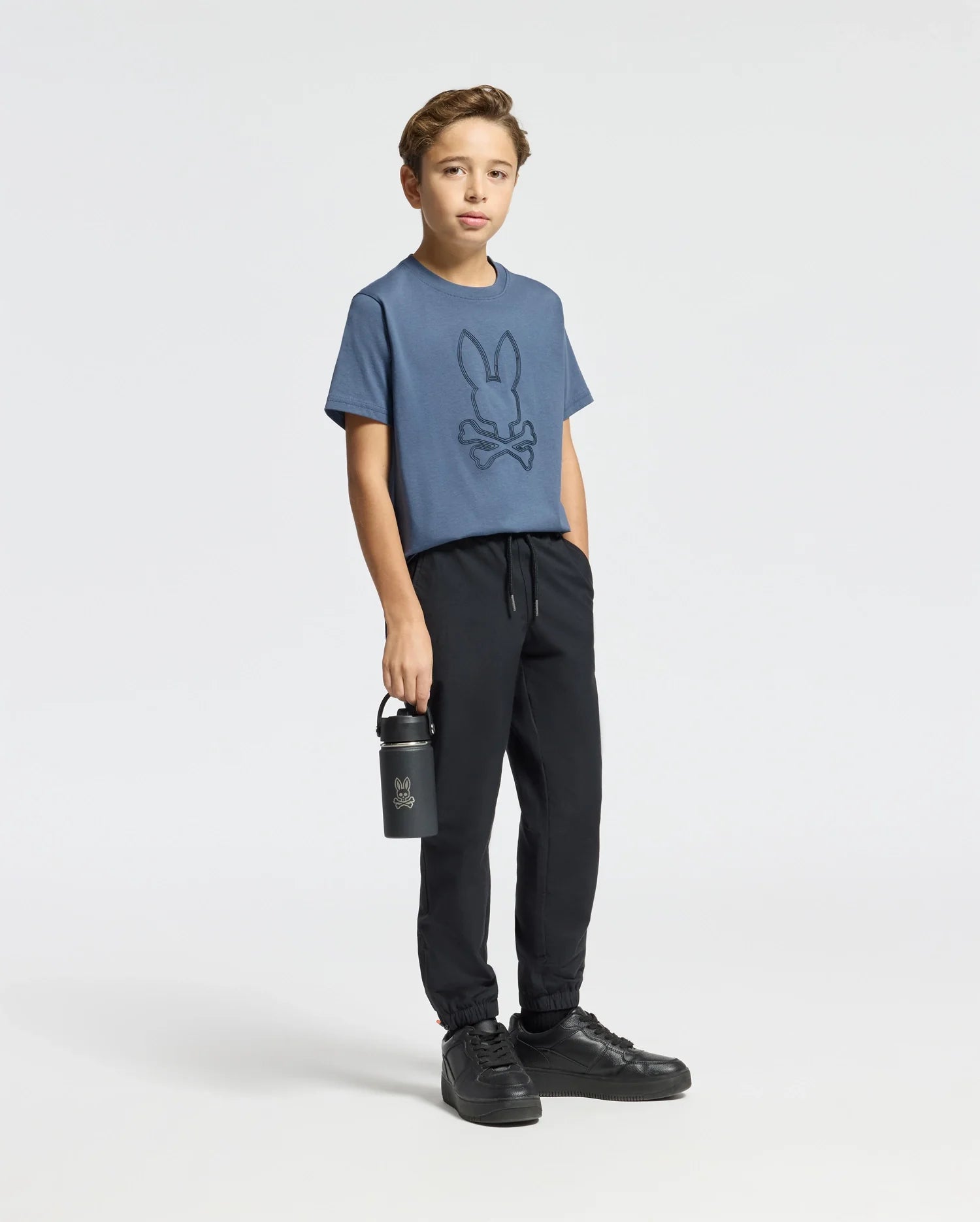 A boy stands against a plain background wearing the Psycho Bunny KIDS MONARC FRONT BUNNY GRAPHIC TEE in blue, crafted from soft Pima cotton and featuring the Monarc Front Bunny logo. He's styled with black jogger pants and sneakers, holding a matching black water bottle with the logo while his other hand rests casually in his pocket.